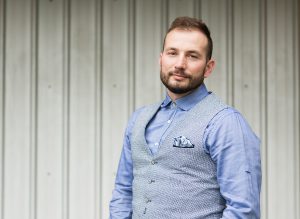 Zack Tica, wearing a blue shirt with a grey vest. Standing infront of a metal cladded wall.